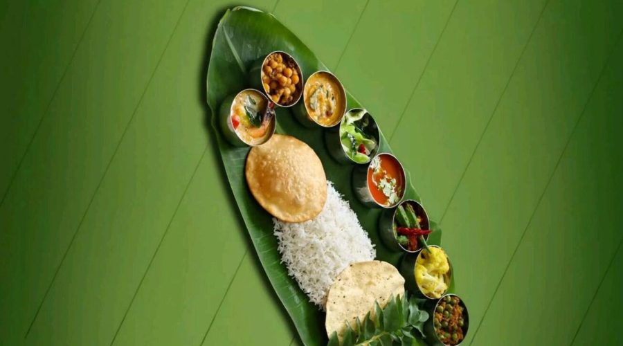A South Indian meal served on a banana leaf with rice, papad, puri, and multiple small bowls of curries, chutneys, and vegetables.